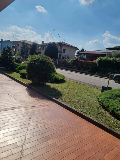 Residential street view with paved terracotta pathway, green hedges, and multi-story houses under blue sky